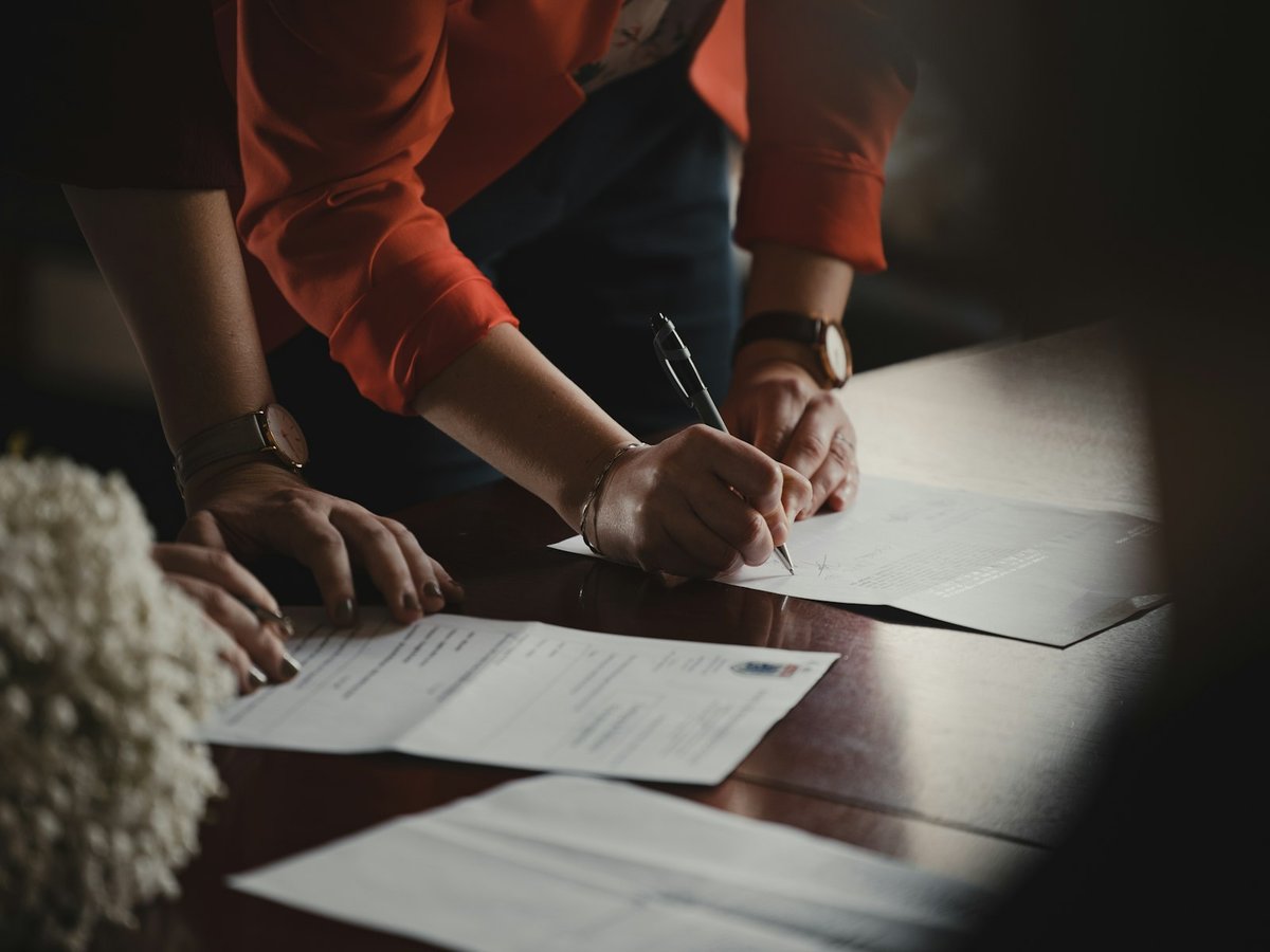 Person in an orange shirt signing papers at a wooden table beside another person